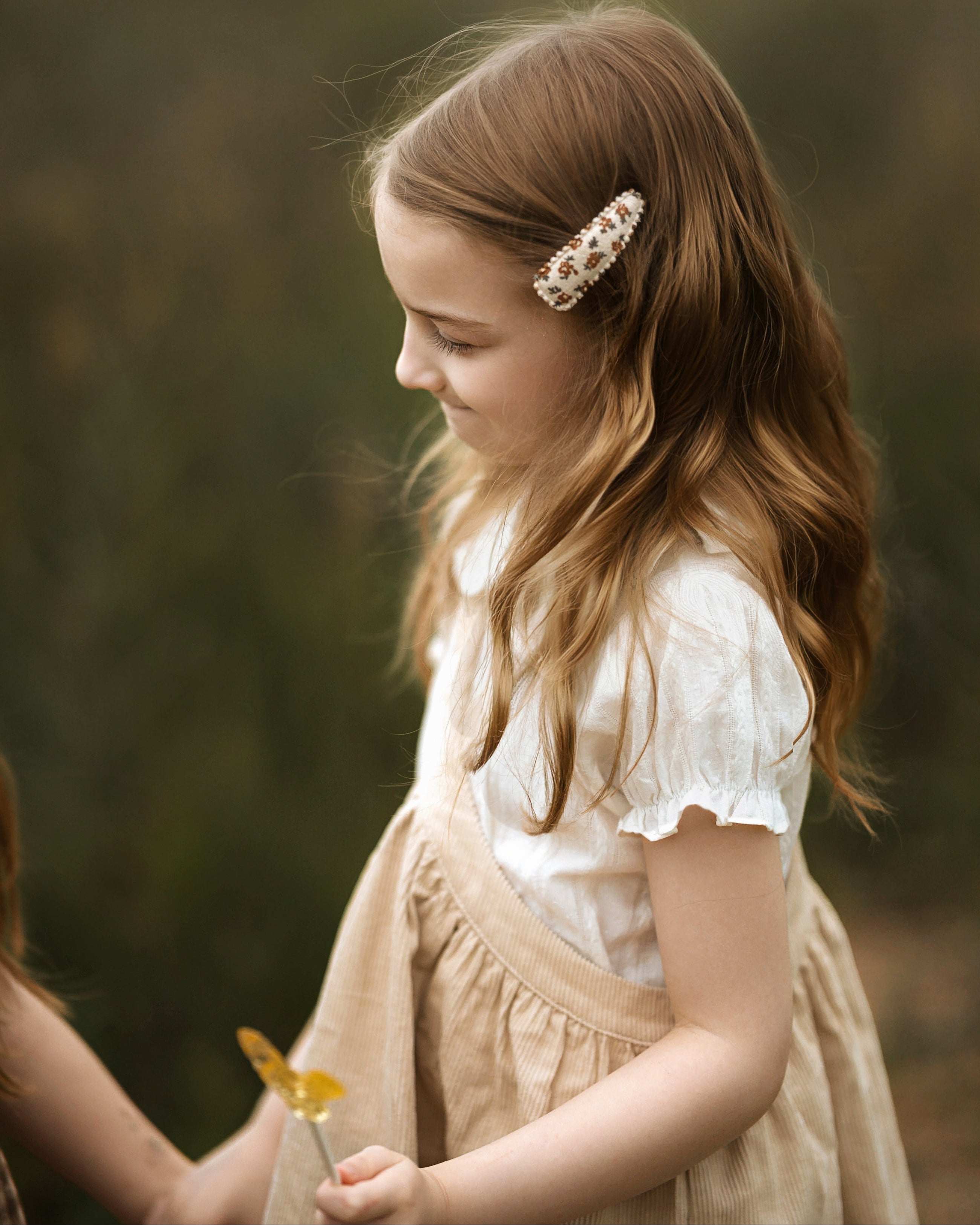A young girl with long light brown hair wearing a white ruffled blouse and beige pinafore dress, holding a yellow flower. She is wearing a decorative pearl and gold barrette hair clip.