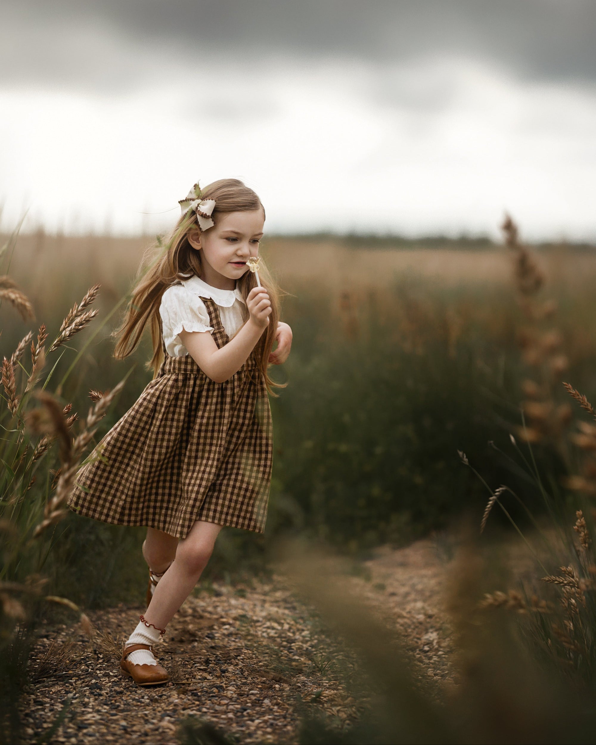 A young girl with a flower hair clip stands on a dirt path in a grassy field, wearing a brown and beige gingham pinafore dress over a white short-sleeve top.