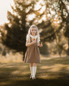 A young girl stands in a sunlit park wearing a brown and beige gingham pinafore dress over a white short-sleeve top, white knee-high socks, and white shoes. She is holding a small orange fruit.