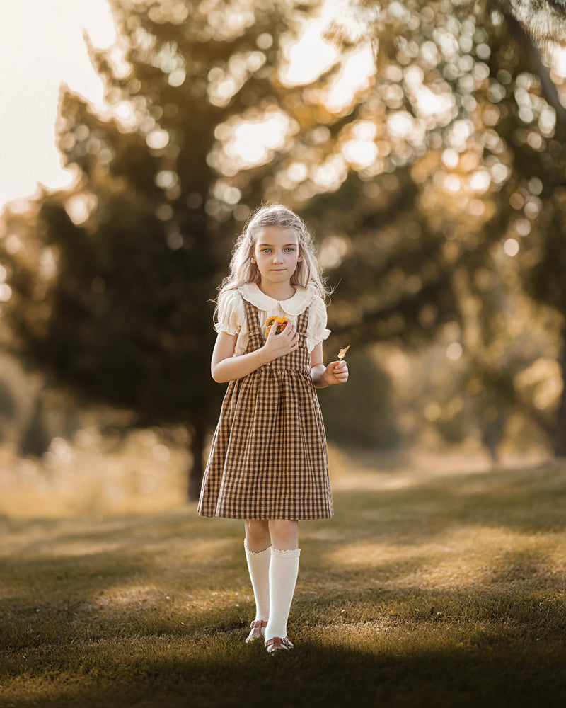 A young girl stands in a sunlit park wearing a brown and beige gingham pinafore dress over a white short-sleeve top, white knee-high socks, and white shoes. She is holding a small orange fruit.