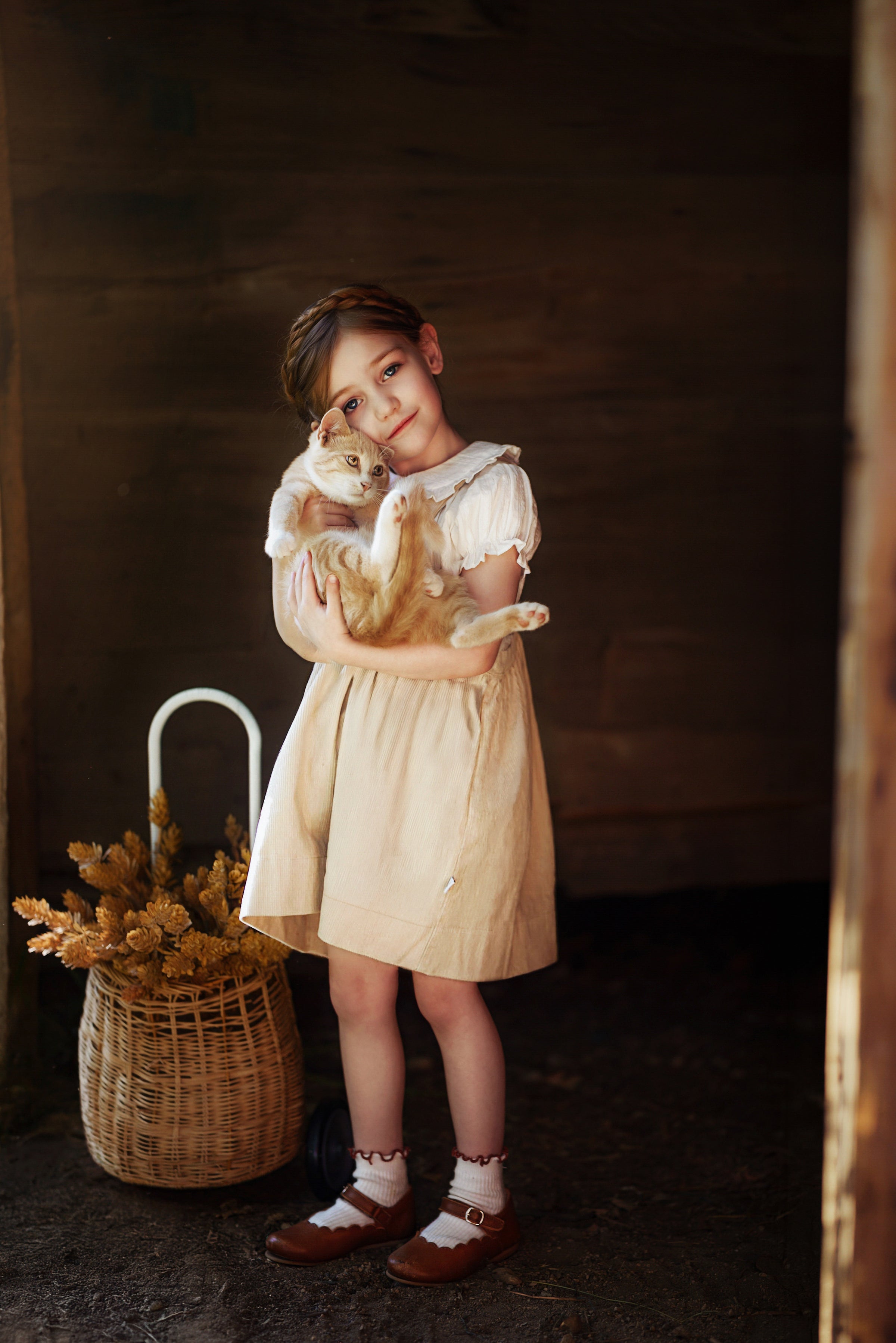 A young girl in a cream-colored vintage-style dress stands in a rustic barn holding a ginger cat, with a wicker basket of dried flowers beside her.