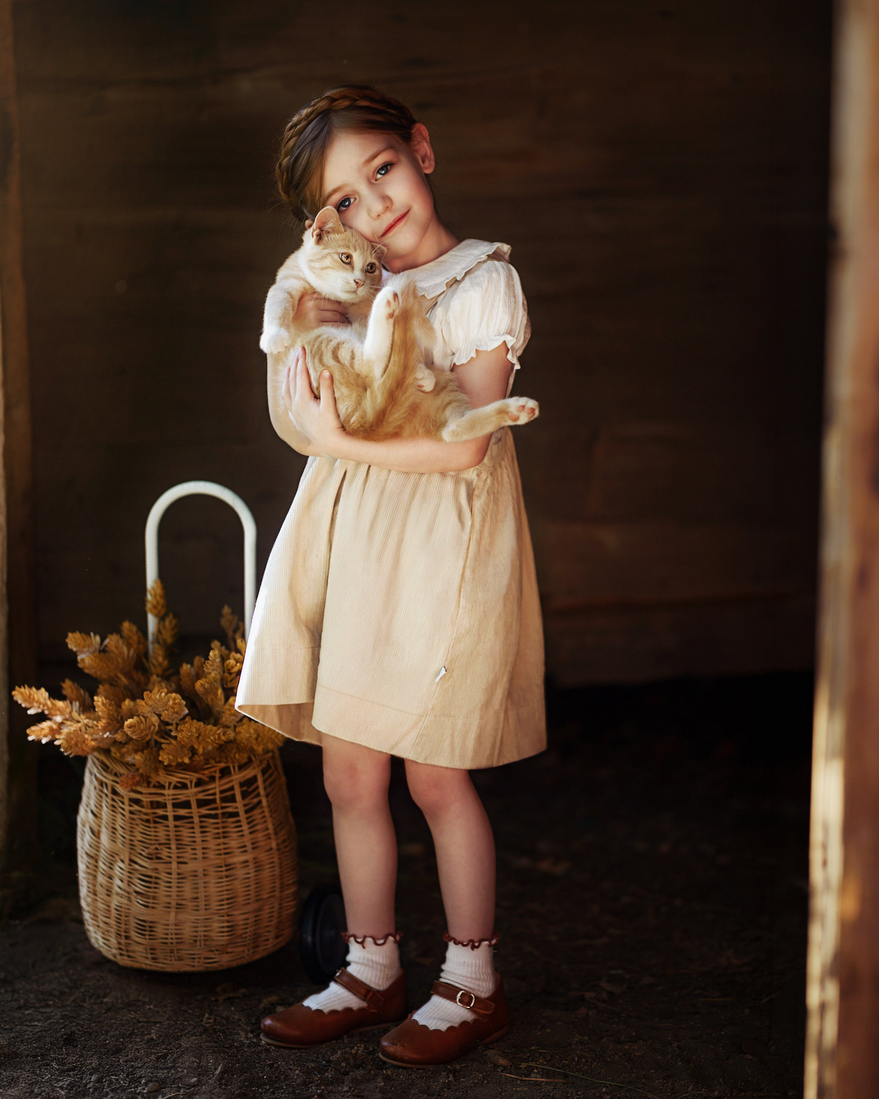 A young girl in a cream-colored dress and brown shoes holds an orange tabby cat in a rustic setting next to a wicker basket with dried flowers.