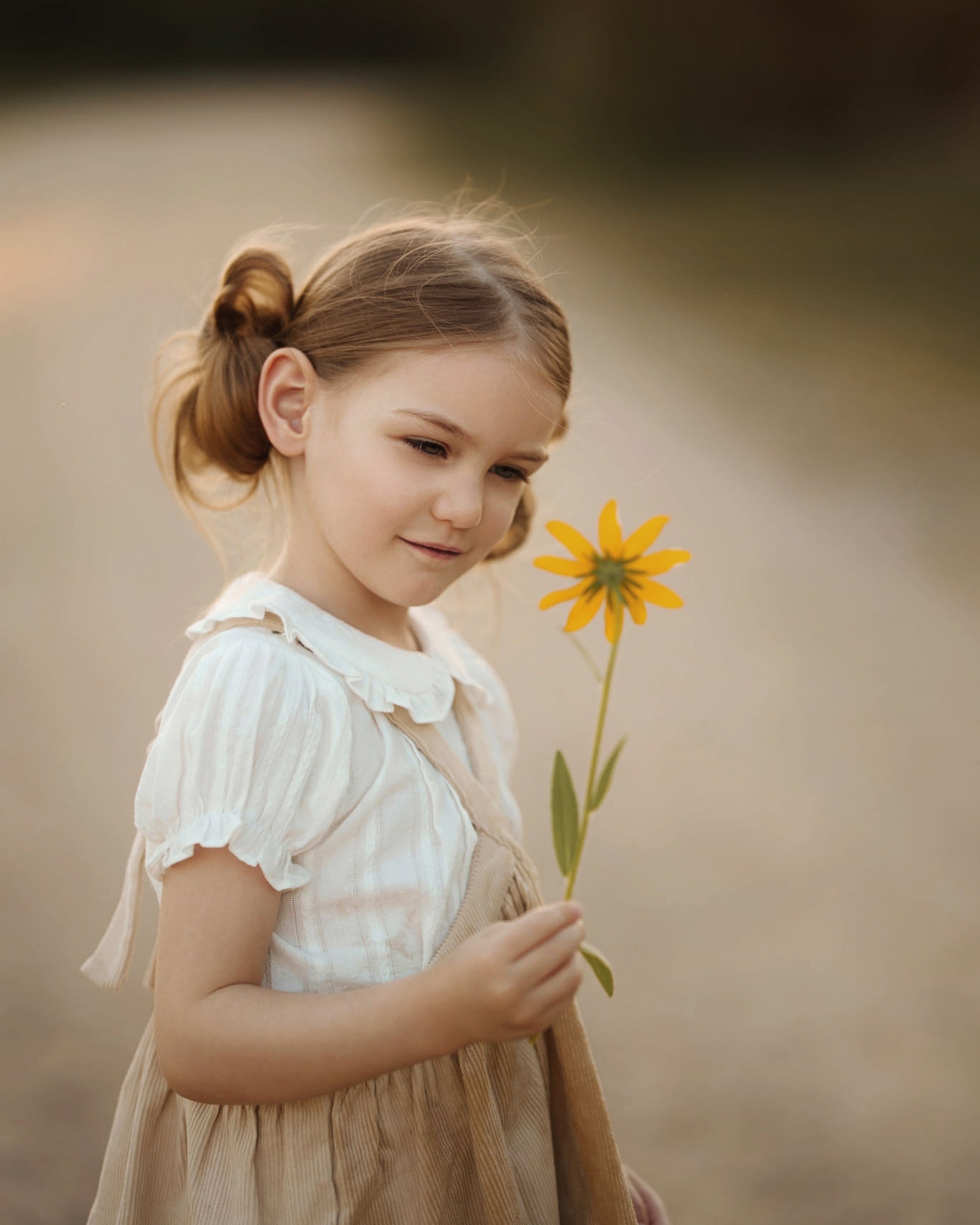 A young girl with her hair in a half-up bun wears a white short-sleeve blouse and a beige pinafore dress, holding a single yellow flower in a soft-focus outdoor setting.