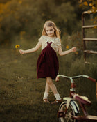 A young girl stands in a grassy outdoor yard holding a small yellow flower, wearing a cream short-sleeve top and a dark burgundy pinafore dress. A vintage-style tricycle is partially visible in the foreground, with trees and a fence in the background under warm golden light.