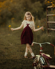 A young girl stands in a grassy outdoor yard holding a small yellow flower, wearing a cream short-sleeve top and a dark burgundy pinafore dress. A vintage-style tricycle is partially visible in the foreground, with trees and a fence in the background under warm golden light.