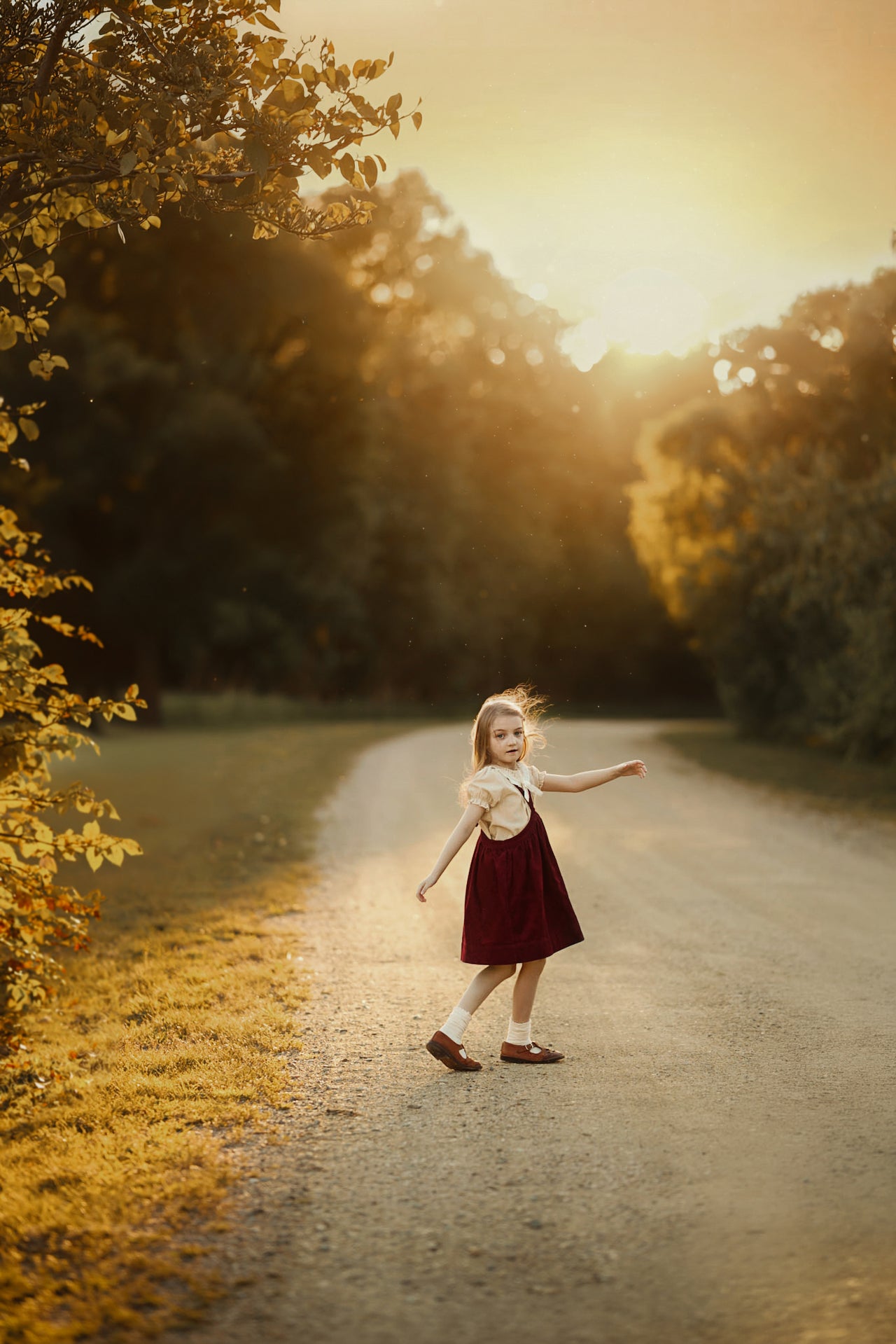 A young girl in a dark red pinafore dress and white blouse stands on a sunlit dirt path surrounded by trees in autumn.