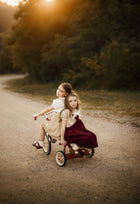 Two young girls riding a small red tricycle on a dirt path outdoors at sunset, with trees in the background. One girl wears a cream dress and the other wears a dark burgundy dress.