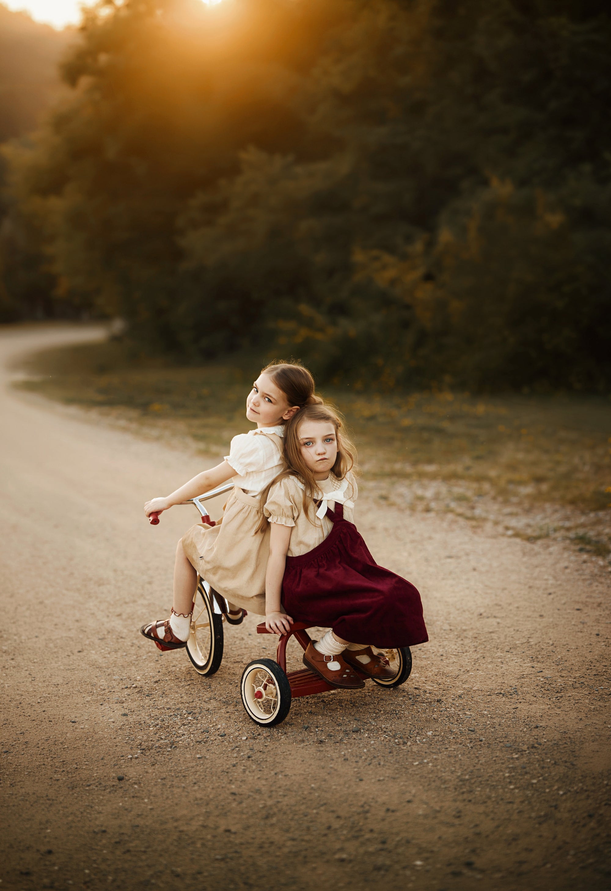 Two young girls riding a small red tricycle on a dirt path outdoors at sunset, with trees in the background. One girl wears a cream dress and the other wears a dark burgundy dress.