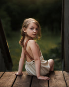 Young girl in a beige dress with brown straps sitting on a wooden platform.