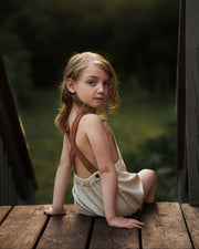 Young girl in a beige dress with brown straps sitting on a wooden platform.