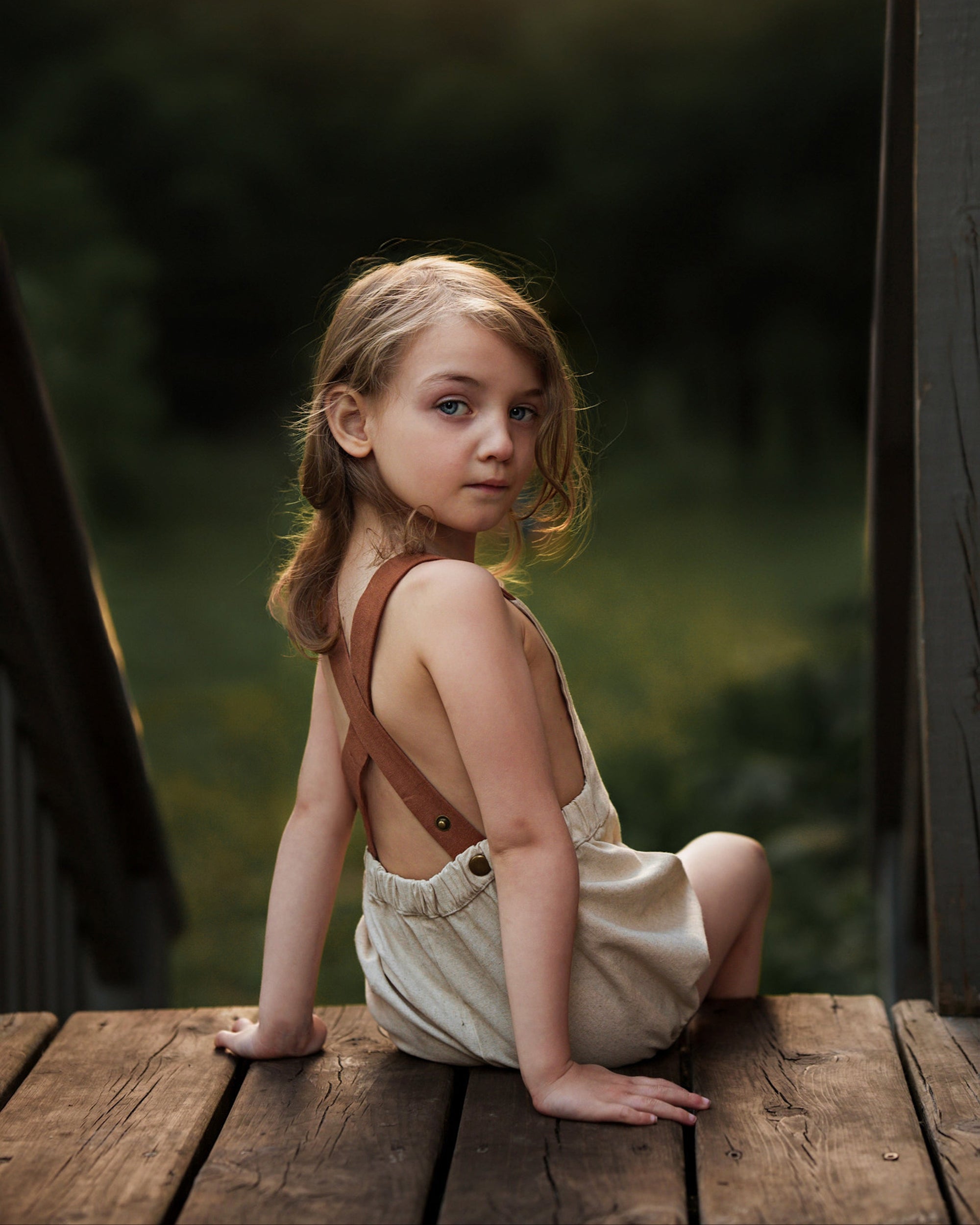 Young girl in a beige dress with brown straps sitting on a wooden platform.