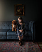 Young girl sitting on a dark gray sofa with a dog, in a dimly lit room.