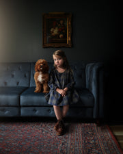 Young girl sitting on a dark gray sofa with a dog, in a dimly lit room.