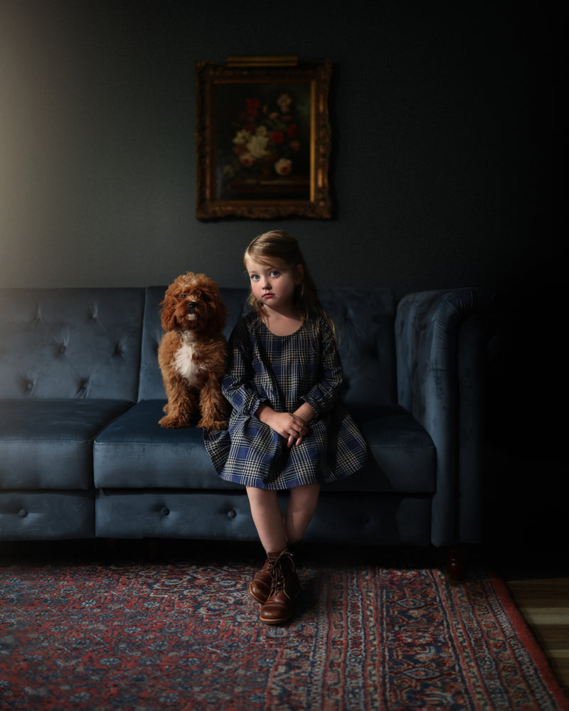 Young girl sitting on a dark gray sofa with a dog, in a dimly lit room.