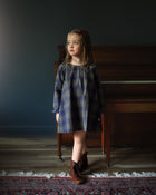 Young girl in a plaid dress standing in a room with a piano.