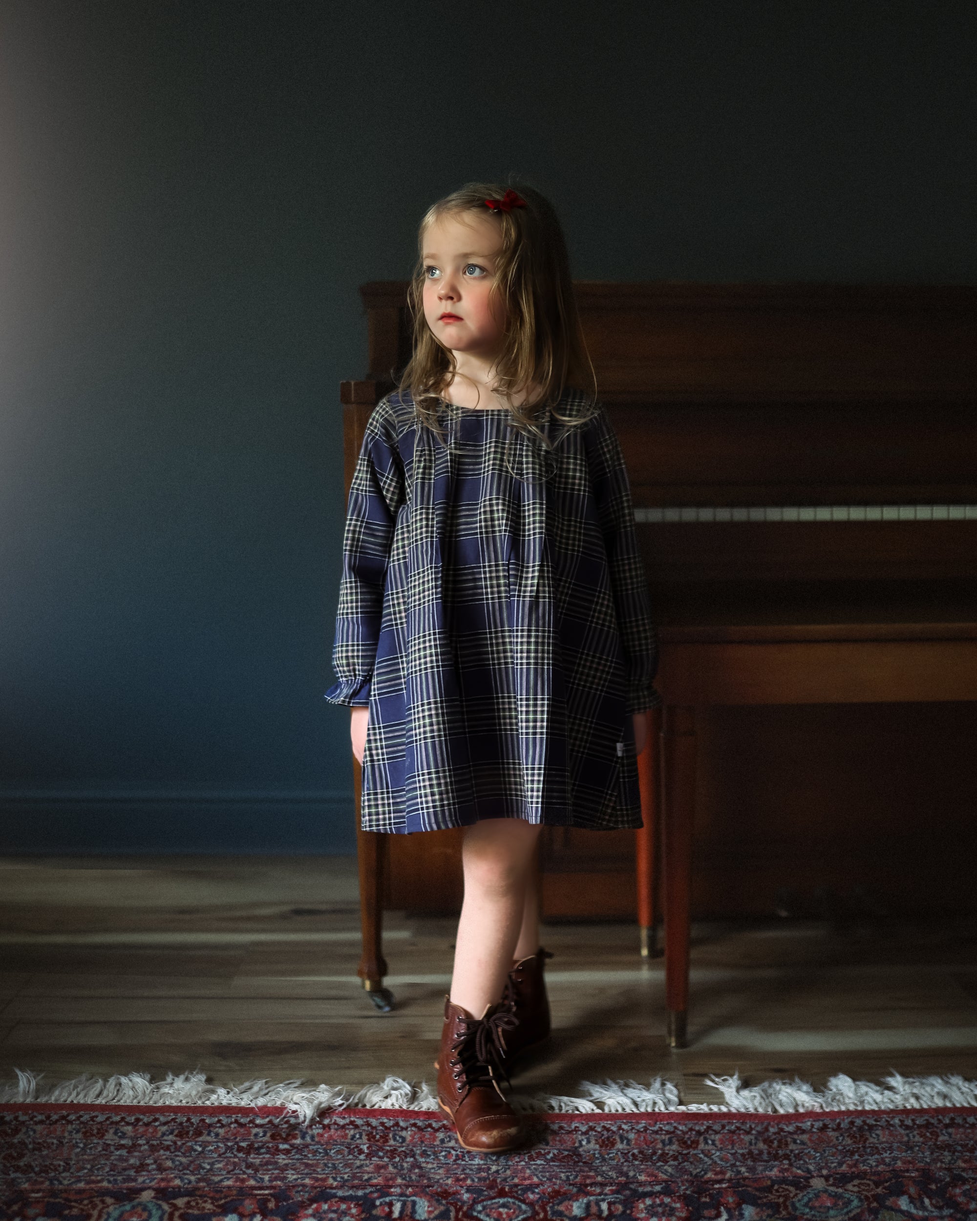 Young girl in a plaid dress standing in a room with a piano.