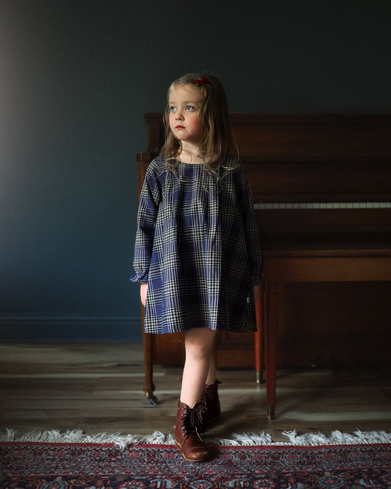 Young girl in a plaid dress standing in a room with a piano.