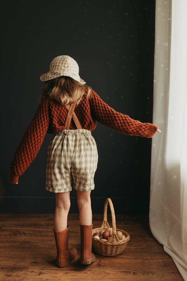 Back view of 5-year-old girl indoors standing next to her Easter eggs, wearing the Grow-With-Me linen Romper in Beige Gingham with brown straps over a cozy sweater, paired with brown rain boots.