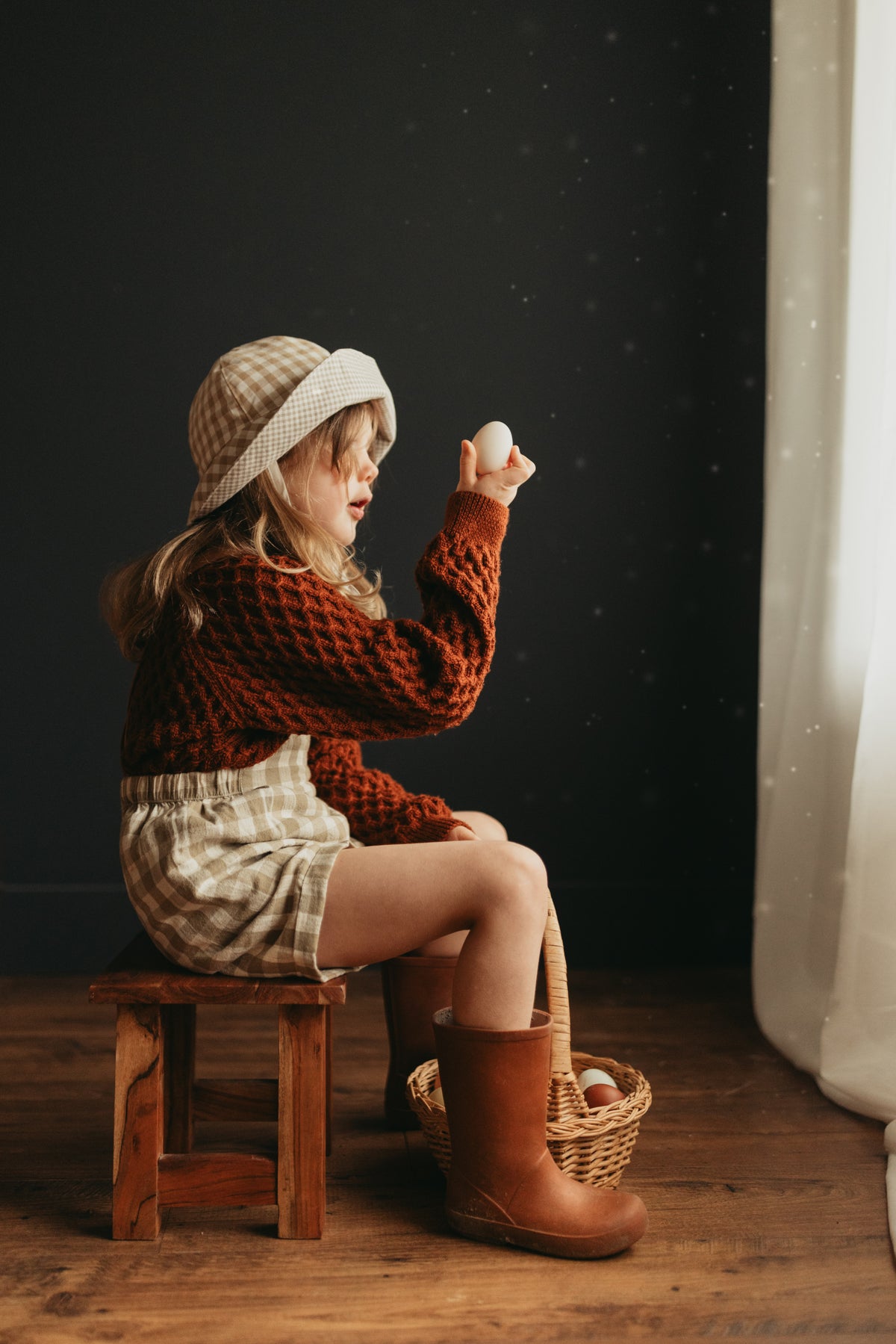 5-year-old girl sitting indoors inspecting her Easter eggs, wearing the Grow-With-Me Romper in Beige Gingham over a cozy sweater, paired with colorful rain boots, highlighting her curiosity and the outfit’s playful style in a cozy indoor setting.