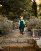A 3-year-old model boy walking outdoors in a classic light blue Henley shirt with white Swiss dots, crafted from natural, sustainable, and high-quality materials. He is paired with spruce corduroy long overalls that enhance the outfit's texture and warmth. This relaxed pose highlights the stylish yet comfortable nature of the ensemble, perfect for everyday adventures.
