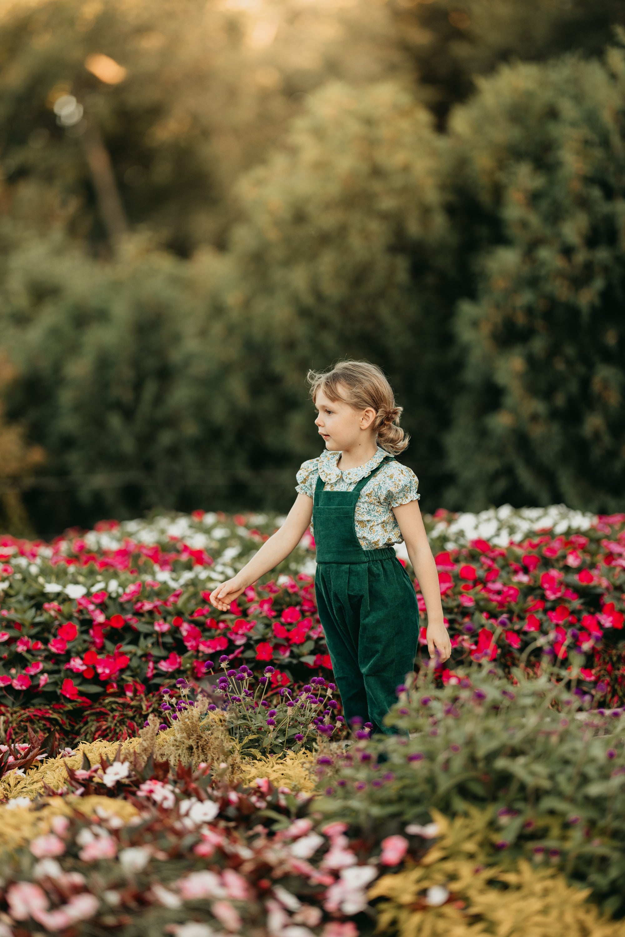 Young girl in green overalls standing among colorful flowers in a garden.