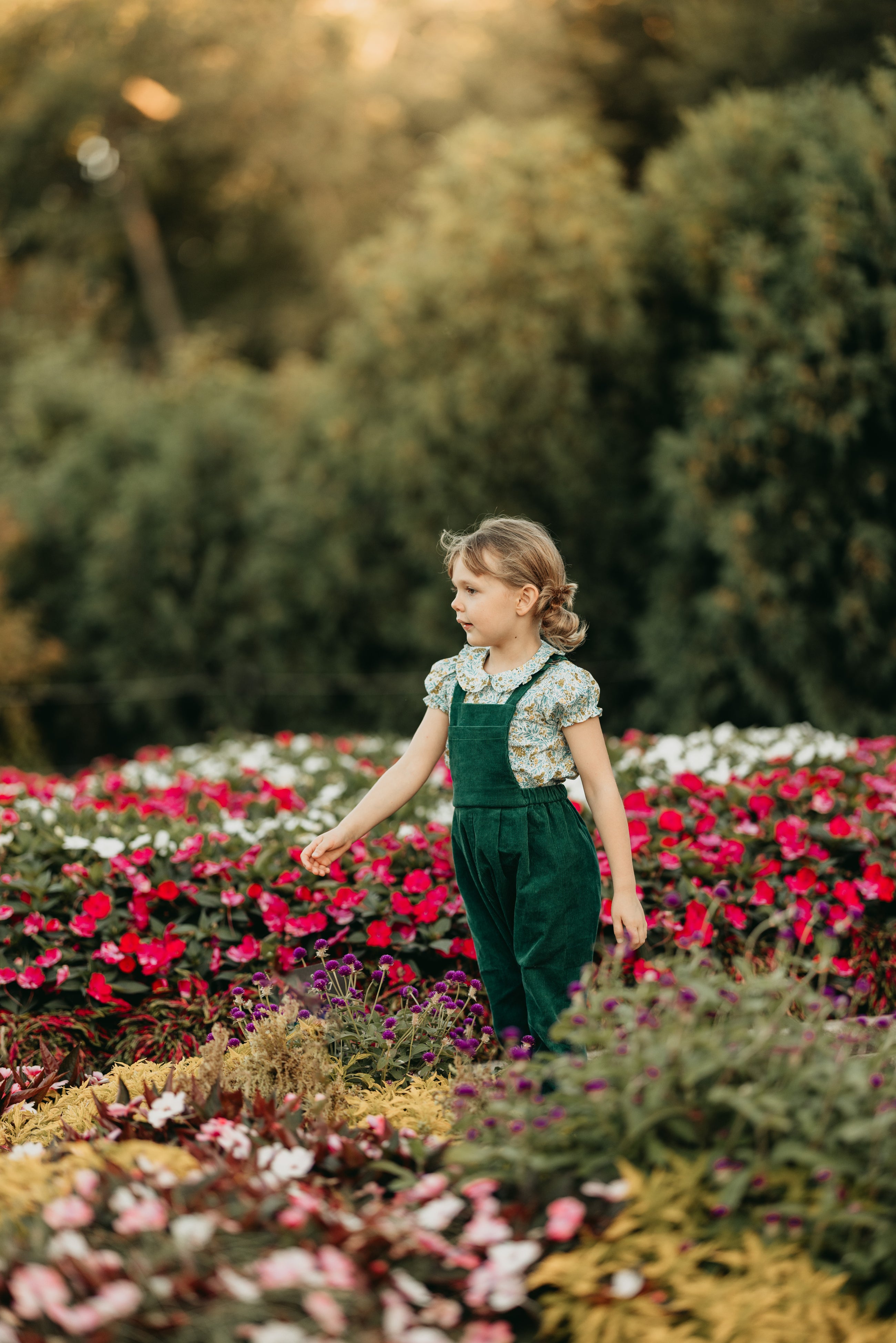 Young girl in green overalls standing among colorful flowers in a garden.