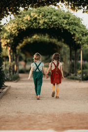 Two girls holding hands and walking away on a path with greenery around.