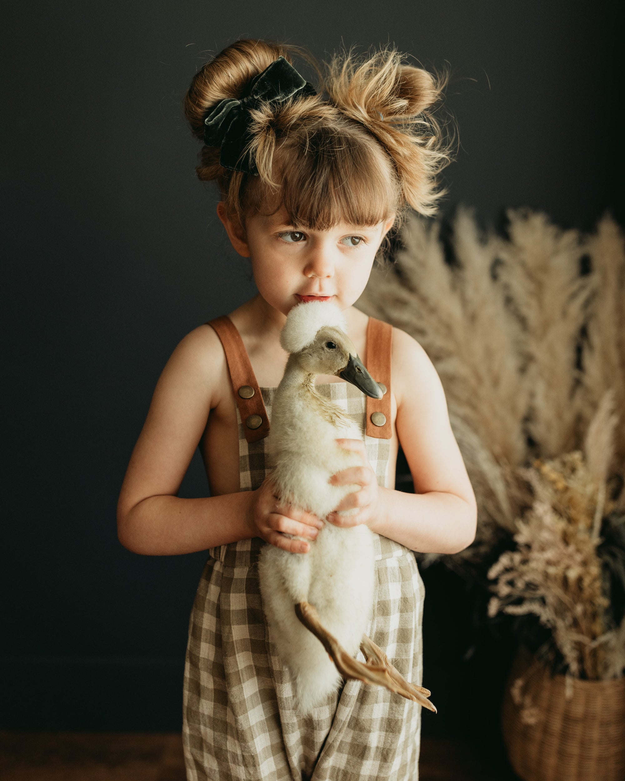 Little 5-year-old girl holding a goose with love, wearing beige gingham linen long overalls featuring a gender-neutral design, breathable fabric for comfort, stylish pleats, durable leather-looking straps, adjustable straps for a perfect fit, and unfoldable leg cuffs designed to grow with her.