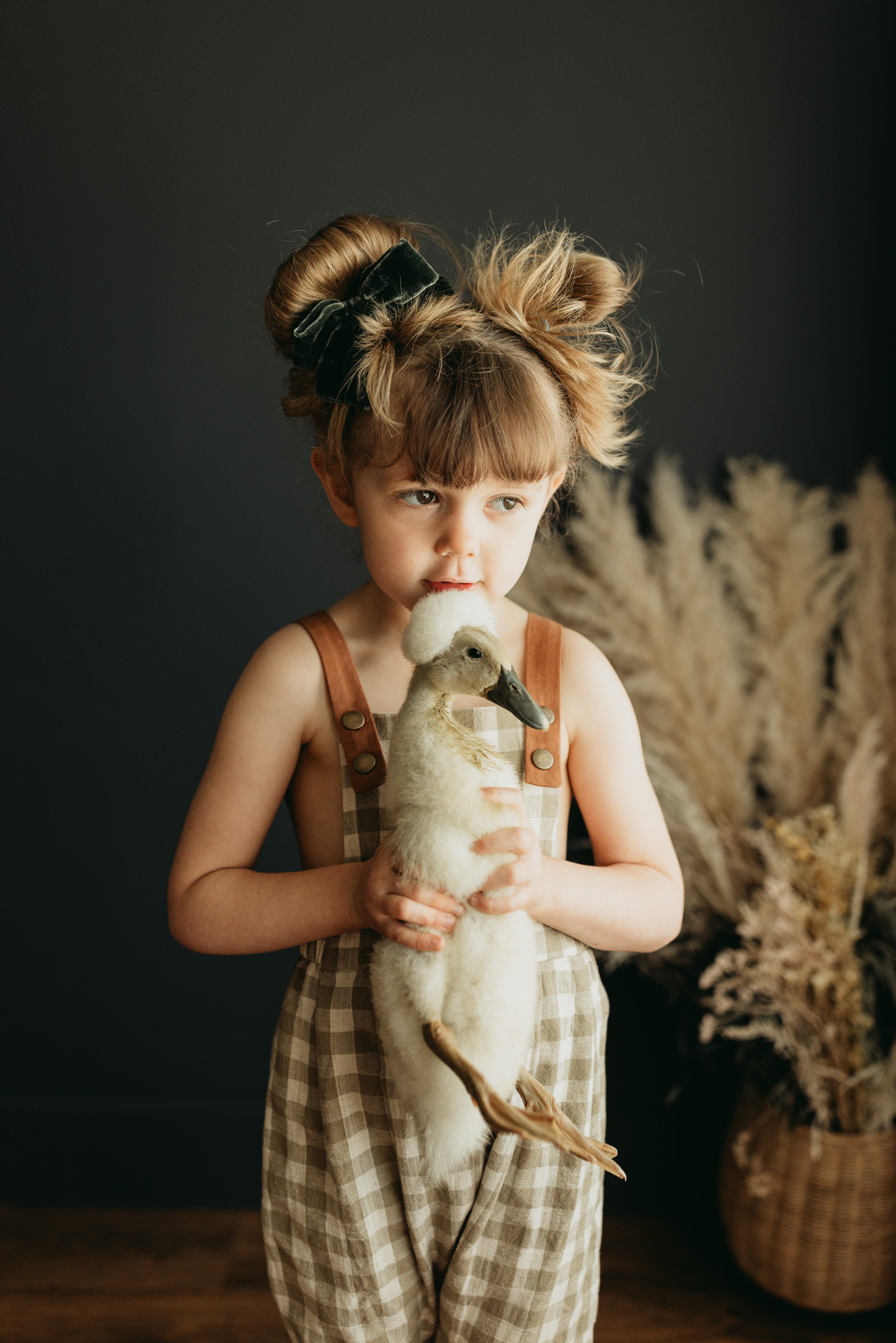 Little 5-year-old girl holding a goose with love, wearing beige gingham linen long overalls featuring a gender-neutral design, breathable fabric for comfort, stylish pleats, durable leather-looking straps, adjustable straps for a perfect fit, and unfoldable leg cuffs designed to grow with her.