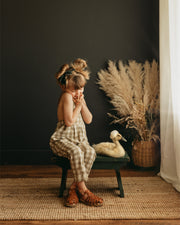 Little 5-year-old girl sitting indoors next to a goose, wearing beige gingham linen long overalls featuring a gender-neutral design, breathable fabric for comfort, stylish pleats, durable leather-looking straps, adjustable straps for a perfect fit, and unfoldable leg cuffs designed to grow with her.