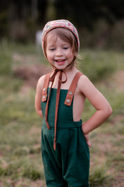 A 3-year-old girl standing and laughing in evergreen long overalls, capturing the joy and playful spirit of young children in stylish clothing.