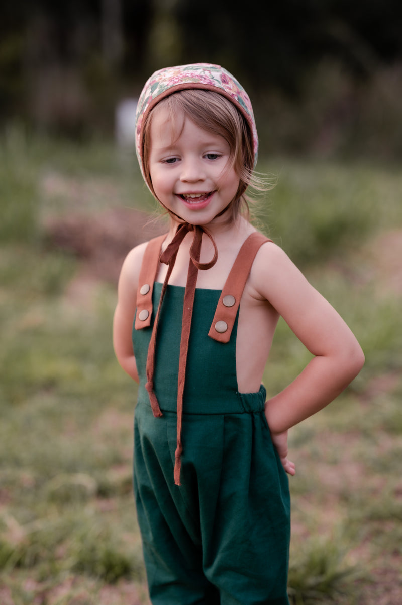 A 3-year-old girl standing and laughing in evergreen long overalls, capturing the joy and playful spirit of young children in stylish clothing.