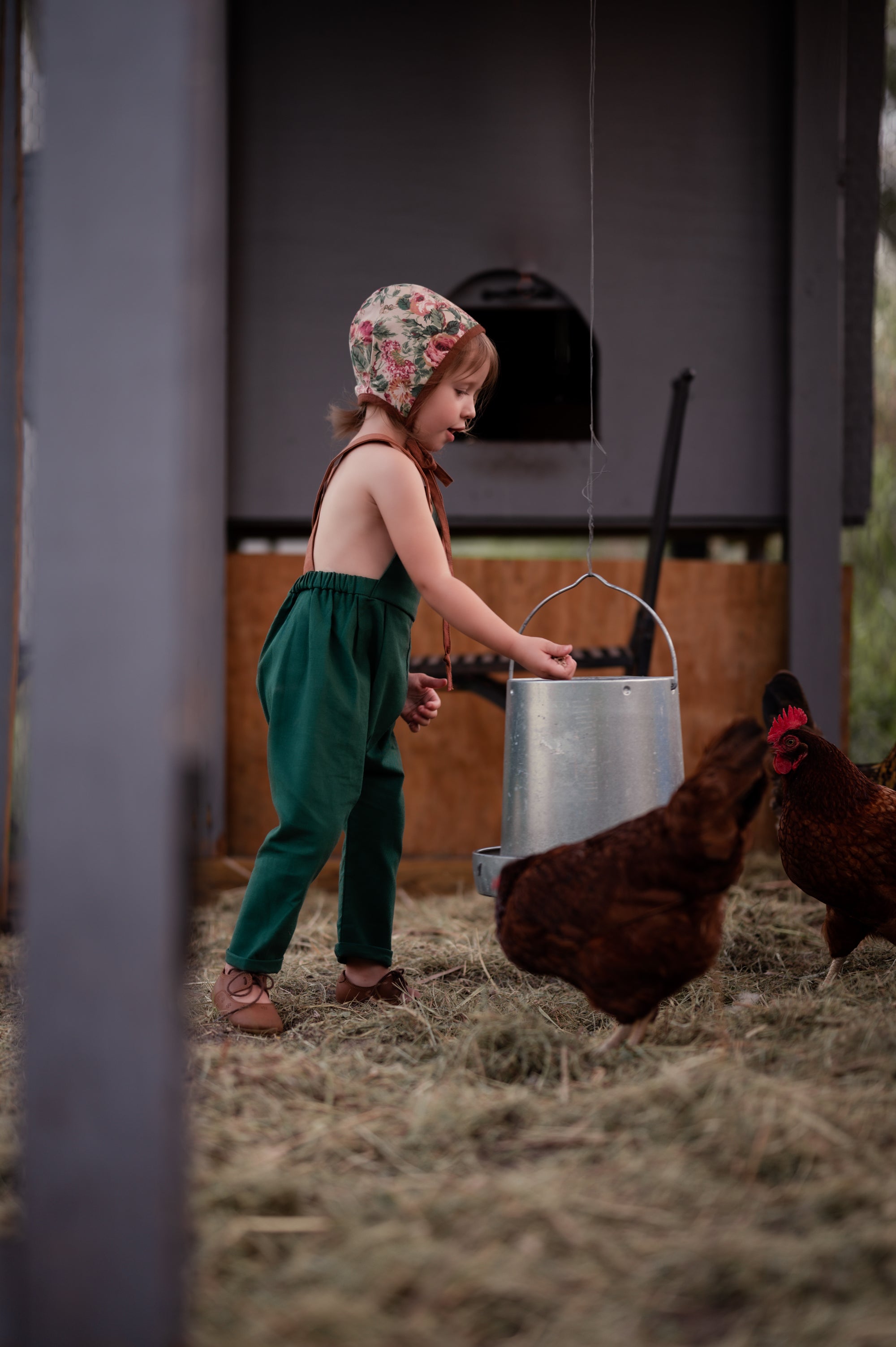 Side view of a 3-year-old girl in the barn wearing evergreen linen long overalls, showcasing the fit and elegant design details that make these overalls a favorite.