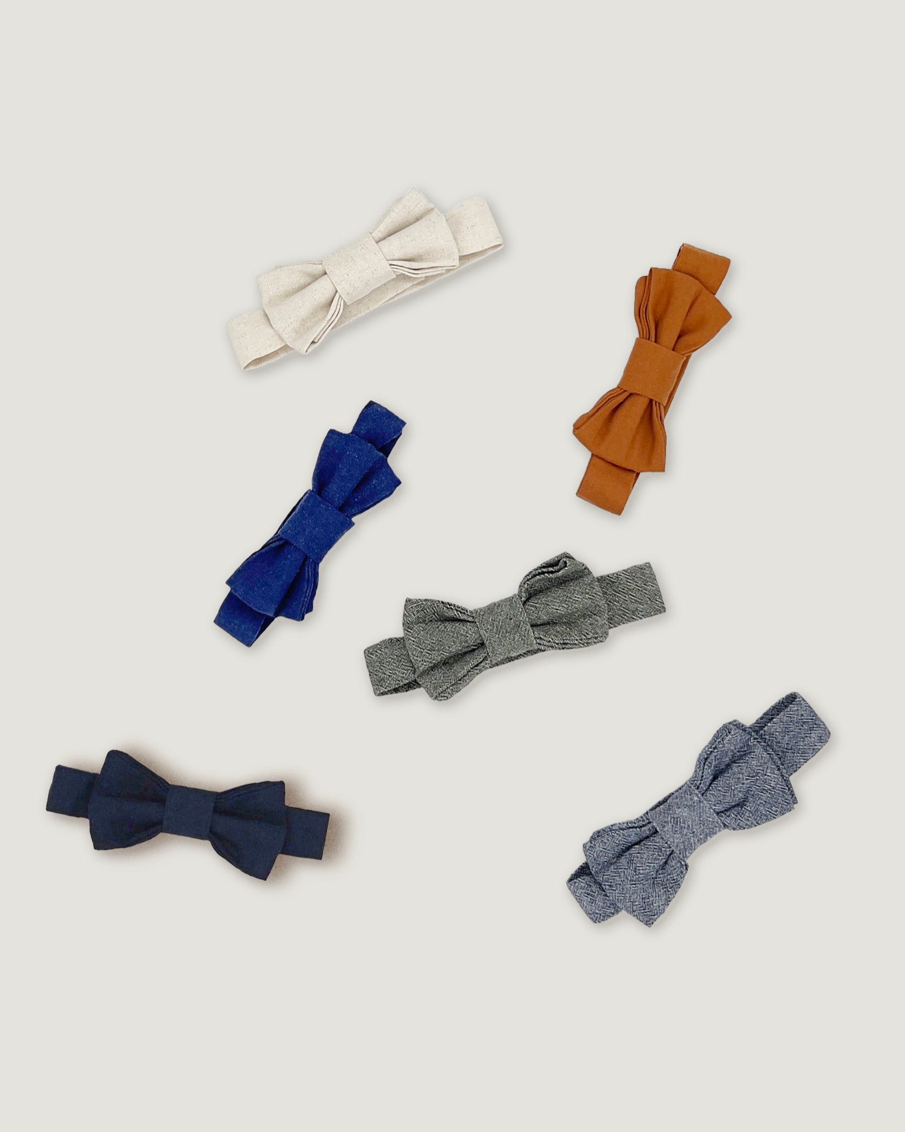 Six fabric bow ties in different colors and textures, including cream, brown, royal blue, heather gray, solid navy, and heather blue, arranged on a light gray background.