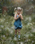 A full view of a little girl in a Blue Jay bubble romper, standing in a sunny field as she blows dandelion seeds. Her joyful expression and the vibrant romper highlight the carefree essence of summer play, surrounded by the beauty of nature.