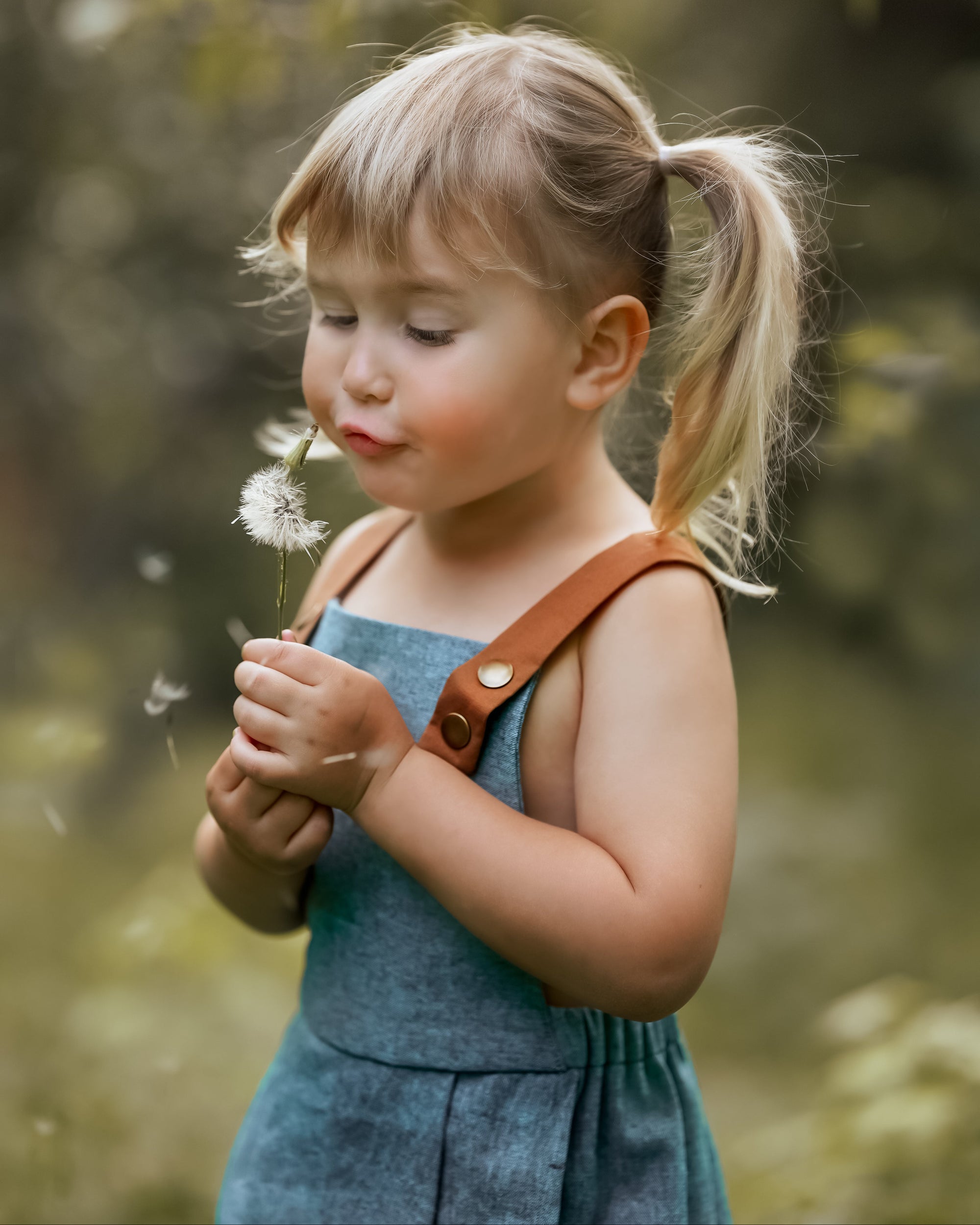 A close-up of a little girl in a Blue Jay bubble romper, joyfully blowing dandelion seeds into the air. Her face is filled with delight, capturing the essence of childhood wonder and the playful spirit of summer.