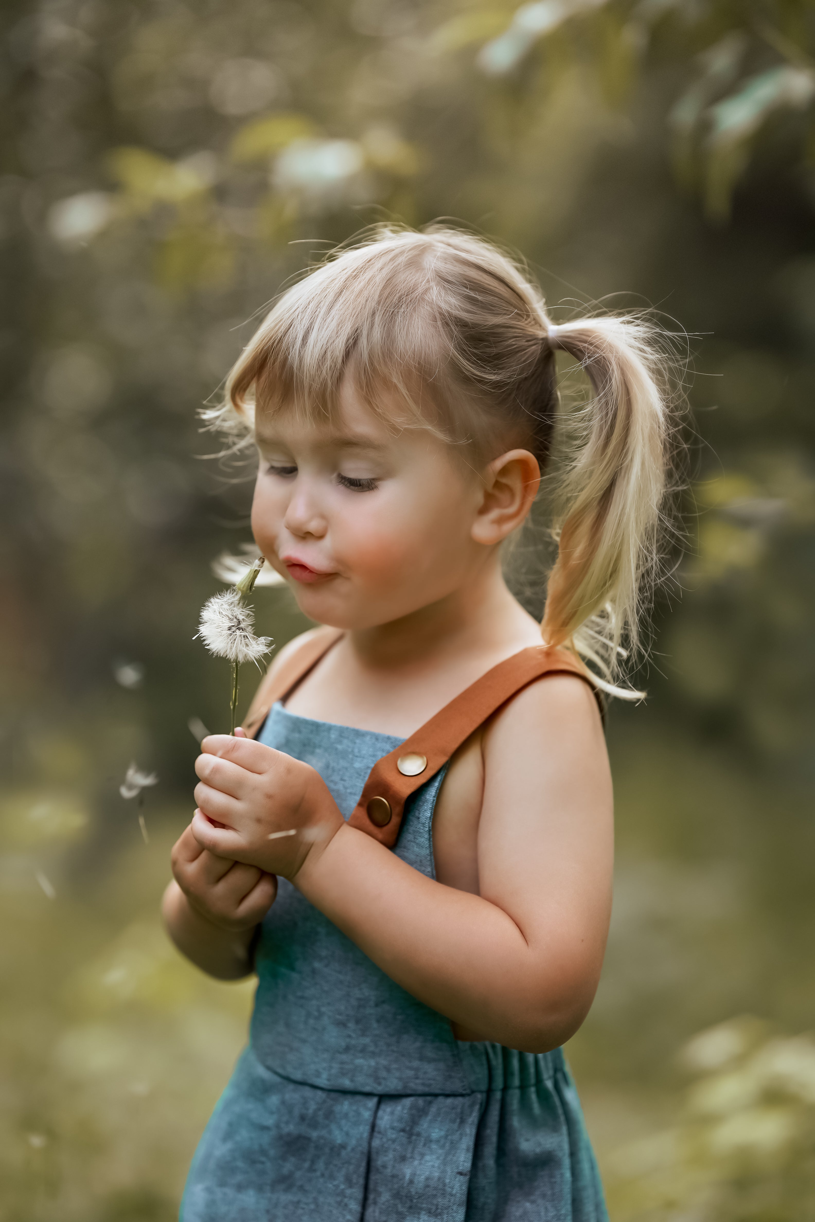 A close-up of a little girl in a Blue Jay bubble romper, joyfully blowing dandelion seeds into the air. Her face is filled with delight, capturing the essence of childhood wonder and the playful spirit of summer.