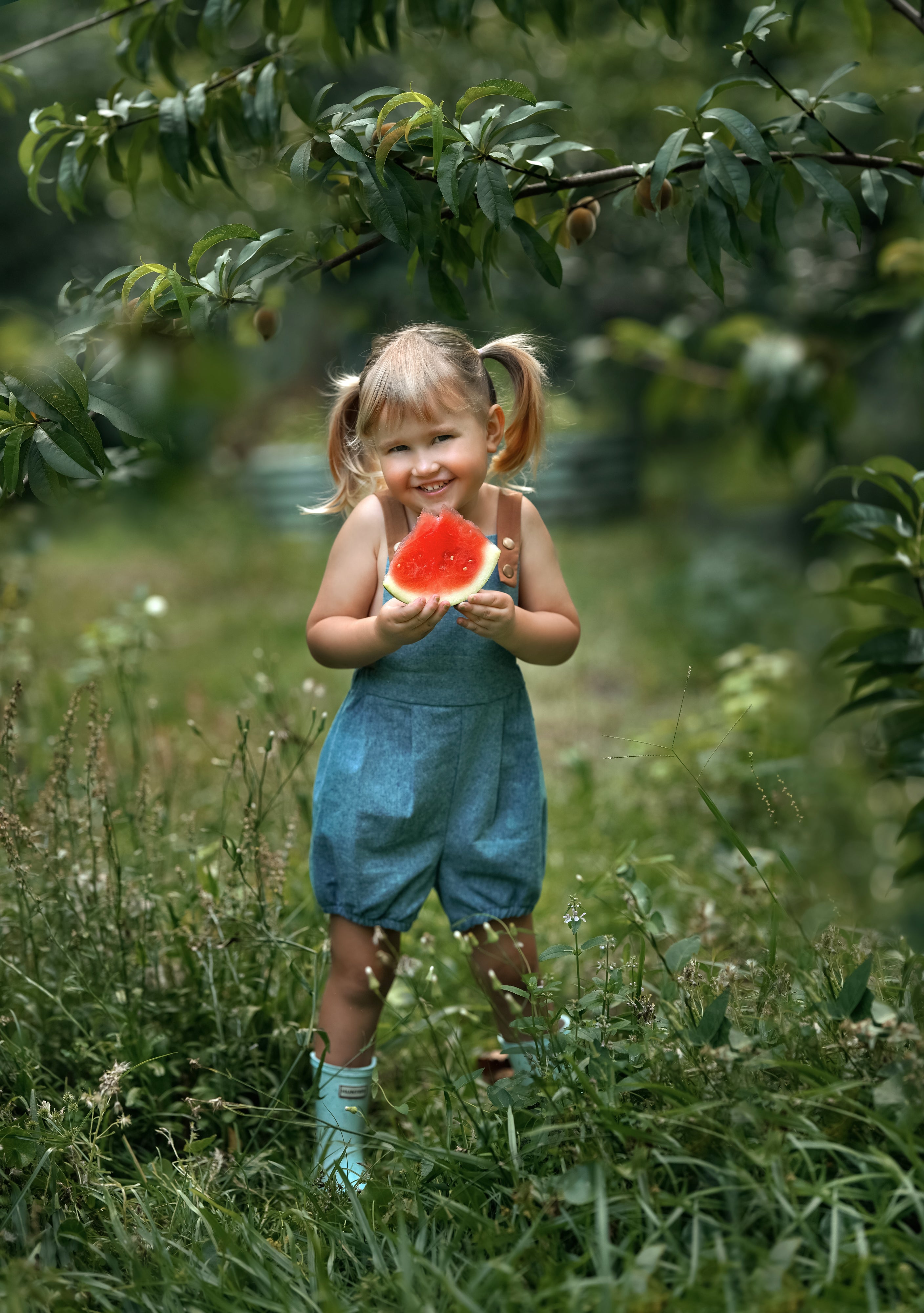 A little girl wearing a Blue Jay bubble romper, smiling brightly while enjoying a slice of watermelon. The romper's adjustable straps and elasticized legs complement her playful demeanor, making it perfect for a summer day.