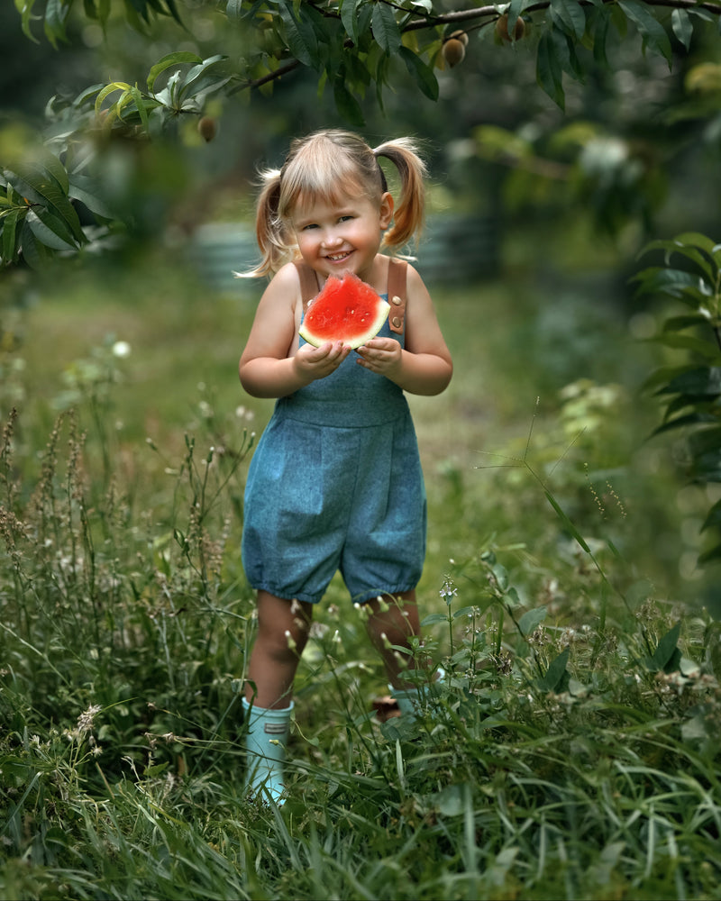 A little girl wearing a Blue Jay bubble romper, smiling brightly while enjoying a slice of watermelon. The romper's adjustable straps and elasticized legs complement her playful demeanor, making it perfect for a summer day.