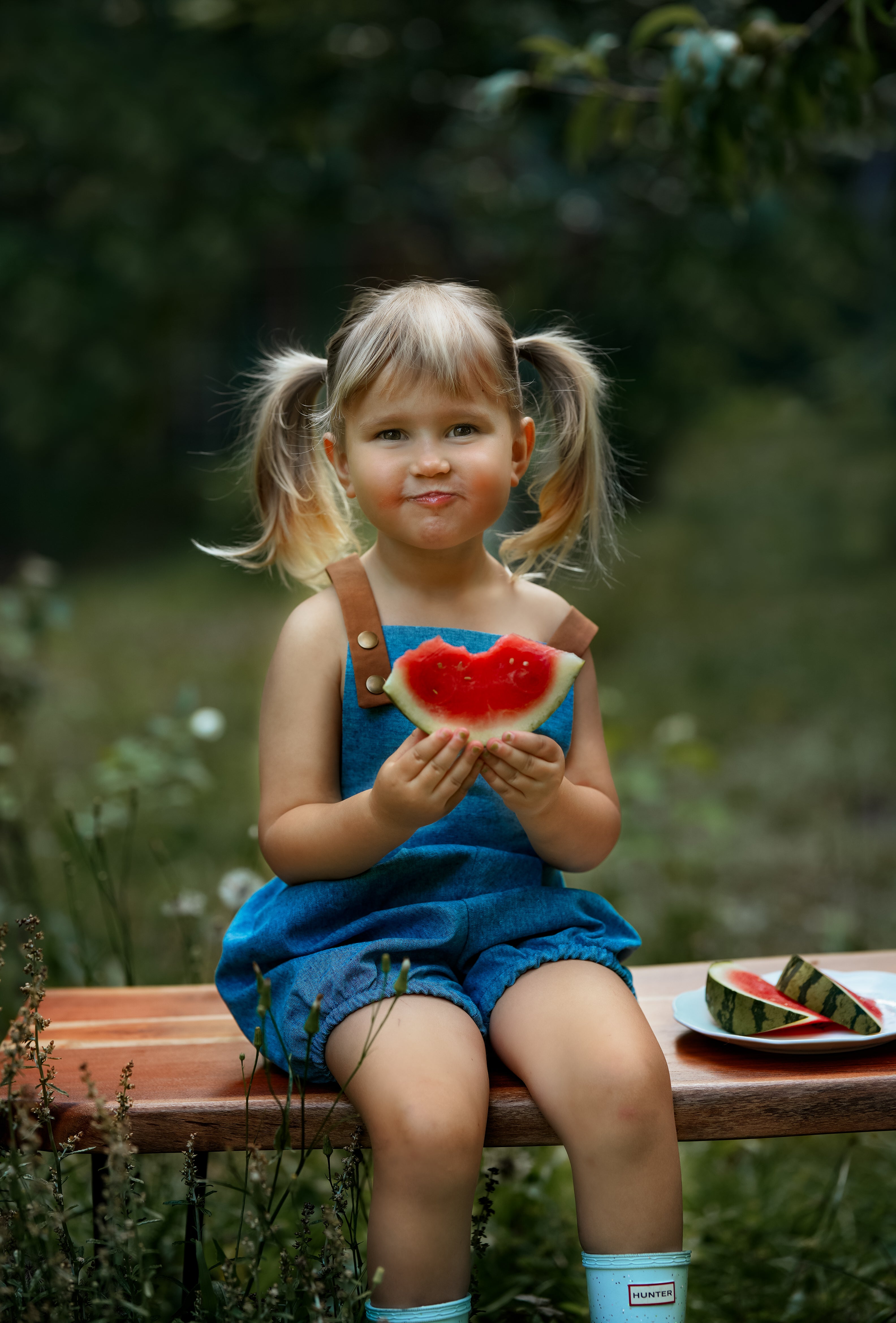 A little girl sitting down in a Blue Jay bubble romper, smiling joyfully while enjoying a slice of watermelon. The romper's adjustable straps and elasticized legs add to her comfort, embodying the carefree spirit of summer.