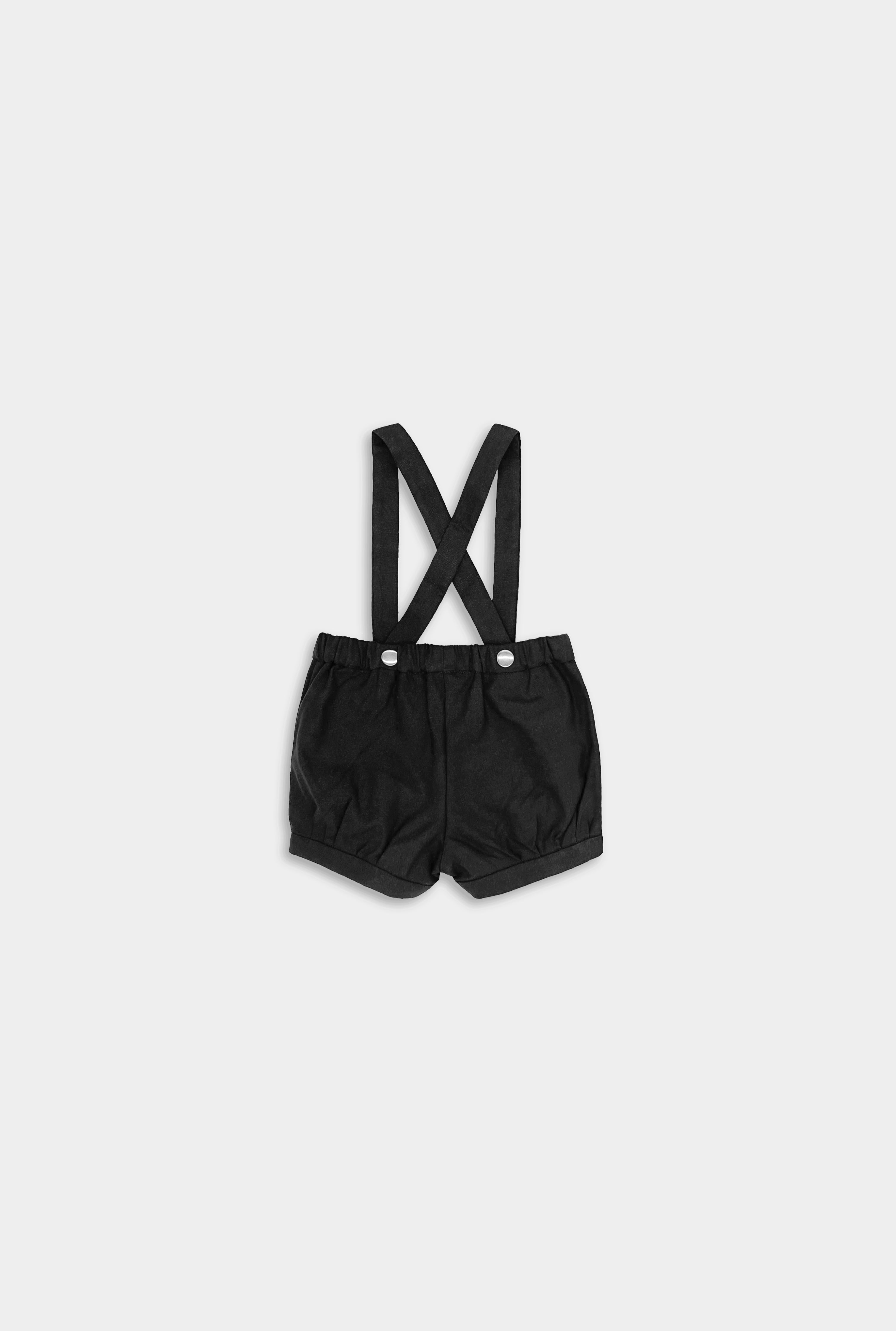 Back view of A pair of black linen suspender shorts for boys featuring adjustable straps for a customized fit, stylish pleats and cuffs for added sophistication, a conveniently positioned label to keep straps up, and versatile strap options with six lengths for flexible styling, allowing them to be worn crossed or straight.