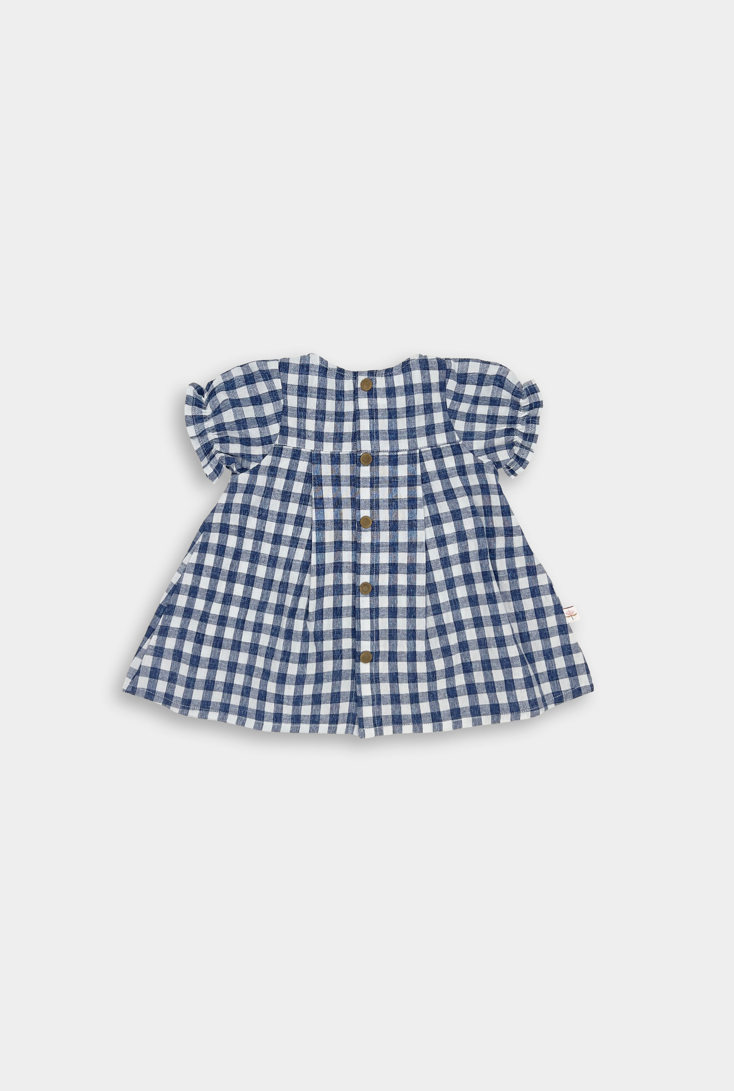 The Blue Gingham linen dress showcases a yoke at the back with pleats and includes secure snaps for easy dressing. This design keeps the overall look simple, allowing the charming gingham pattern to stand out beautifully.