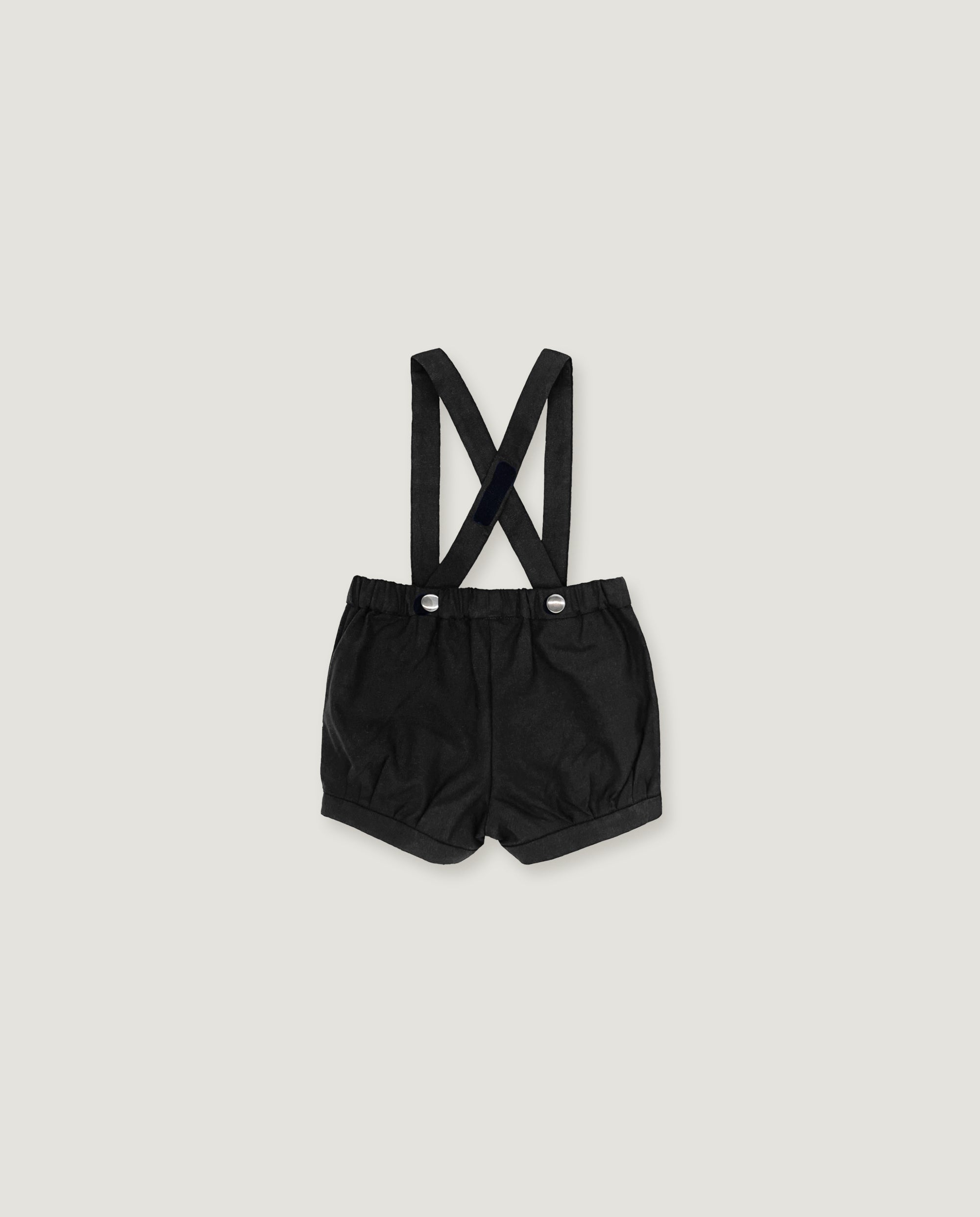 Black baby boy's shorts with adjustable shoulder straps and silver snap buttons, laid flat on a light gray background.