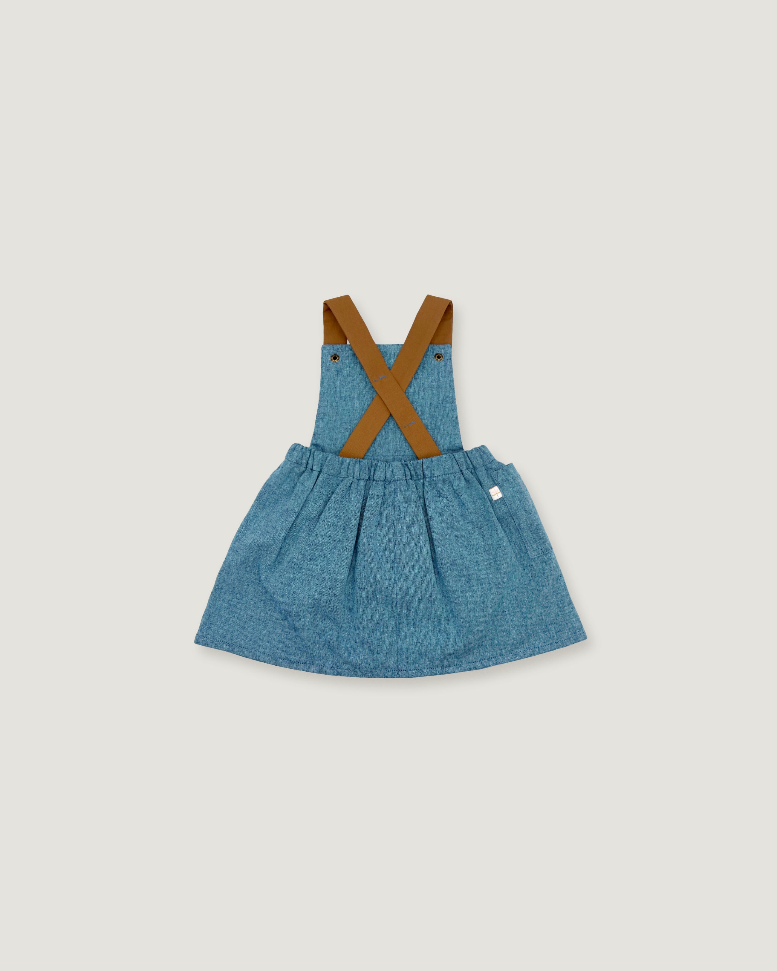 A toddler girl's denim overall dress with brown faux-leather suspenders, shown flat on a light gray background.