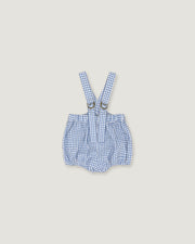 A baby romper with suspenders in a blue and white gingham check pattern, featuring adjustable shoulder straps with metal rings, laid flat on a light beige background.
