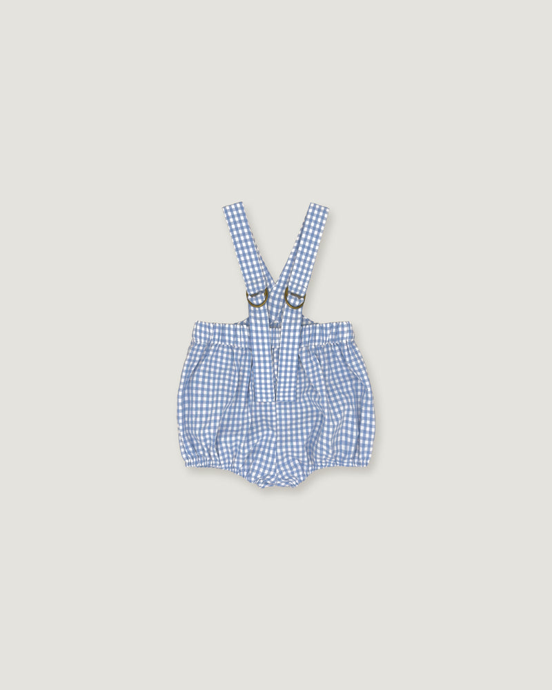 A baby romper with suspenders in a blue and white gingham check pattern, featuring adjustable shoulder straps with metal rings, laid flat on a light beige background.