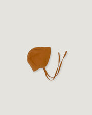 A small brown bonnet-style hat with long ties, photographed flat against a light warm gray background.
