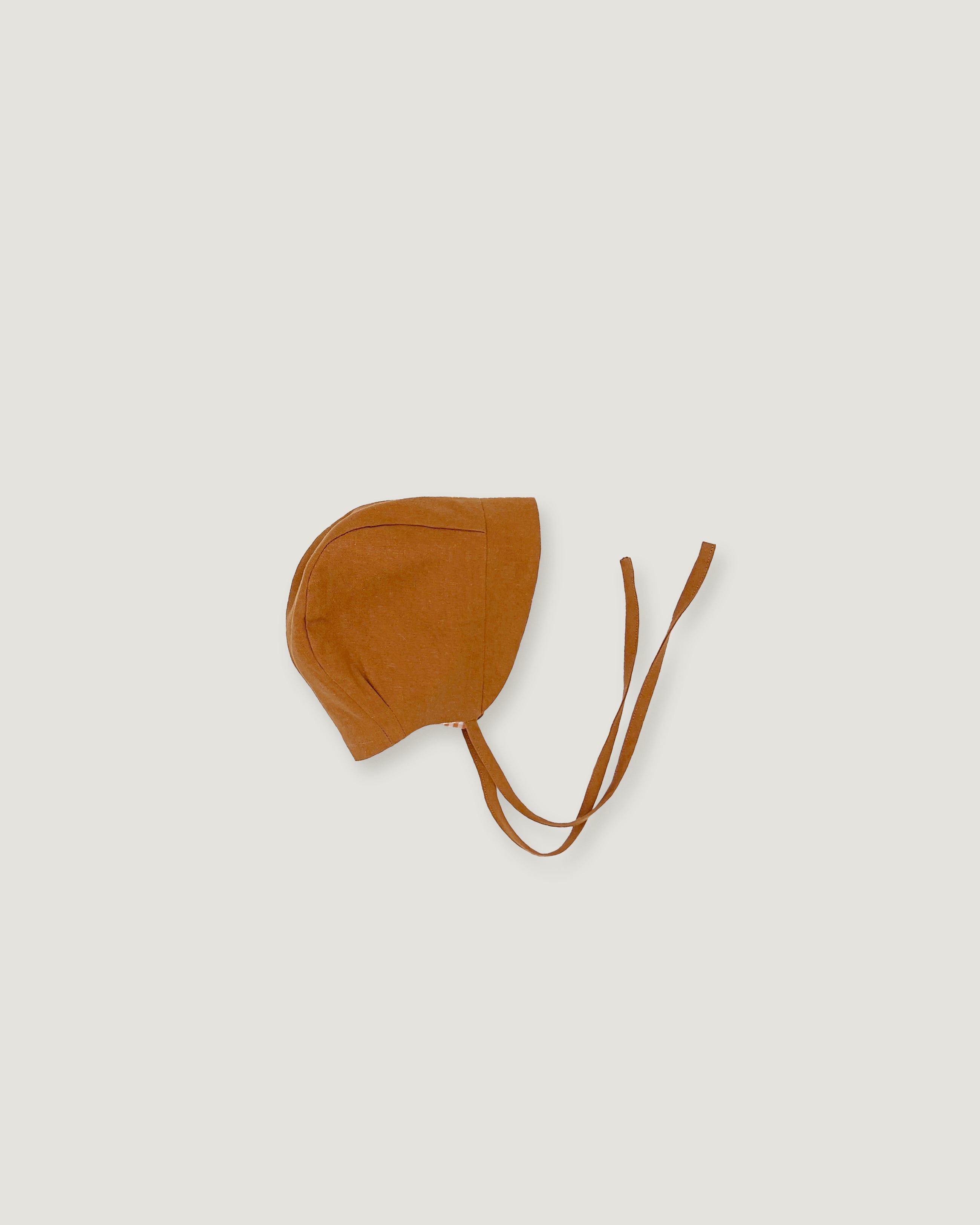A small brown bonnet-style hat with long ties, photographed flat against a light warm gray background.