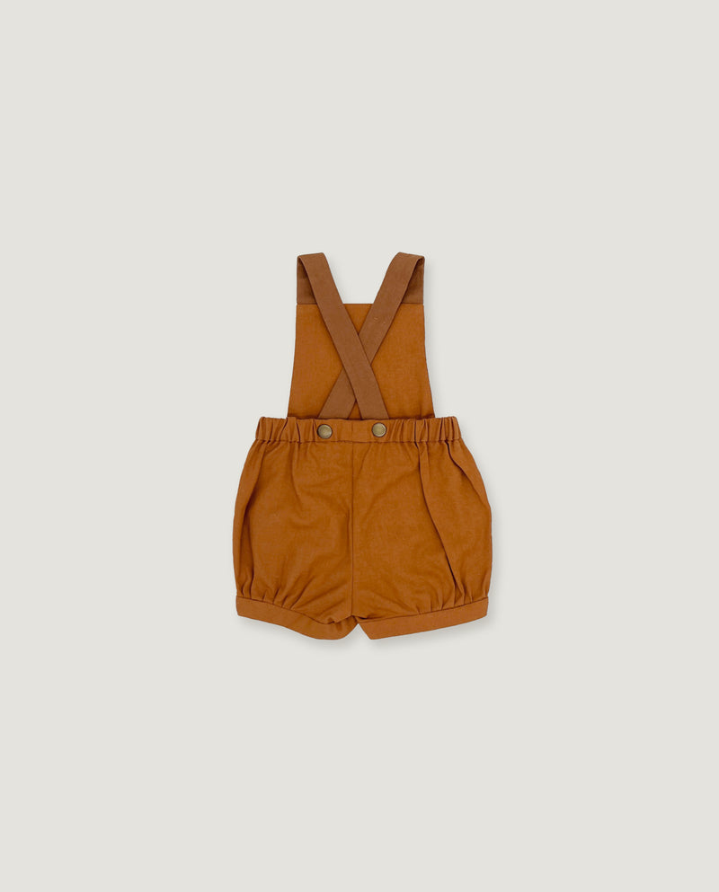 A pair of toddler boy's shortalls in a warm camel brown color, featuring adjustable shoulder straps and a button closure at the waist, displayed flat against a light gray background.