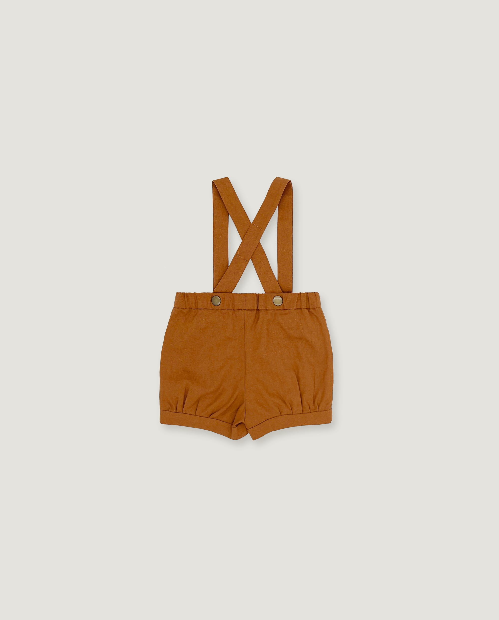 A pair of toddler's short trousers in a warm camel brown color, featuring adjustable shoulder straps that cross over at the back and snap button closures at the waist, displayed flat against a light gray background.